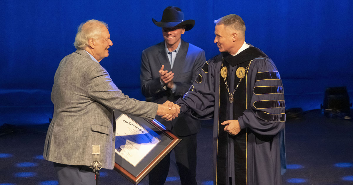 ETSU President Brian Noland shakes hands with Jack Tottle during an award presentation, as Kenny Chesney applauds in the background on stage.