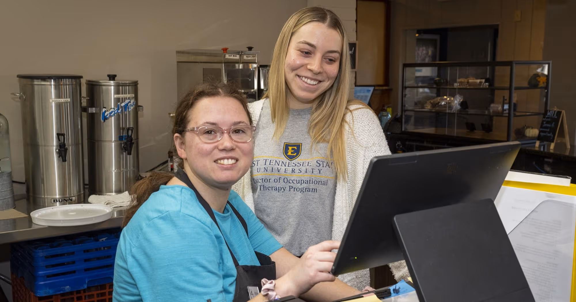 An ETSU student smiles while looking at a computer and posing for a photo with an employee of the Jeremiah Cafe.