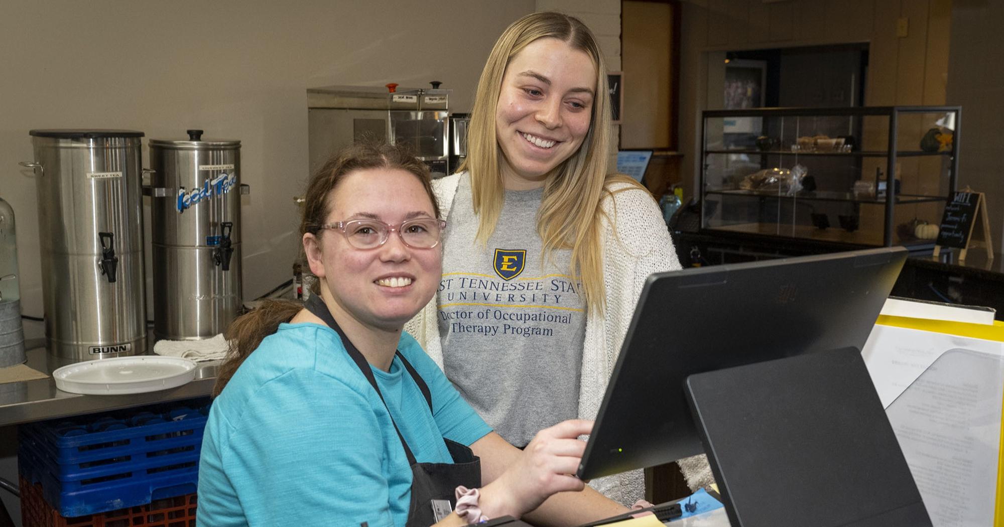 An ETSU student smiles while looking at a computer and posing for a photo with an employee of the Jeremiah Cafe.