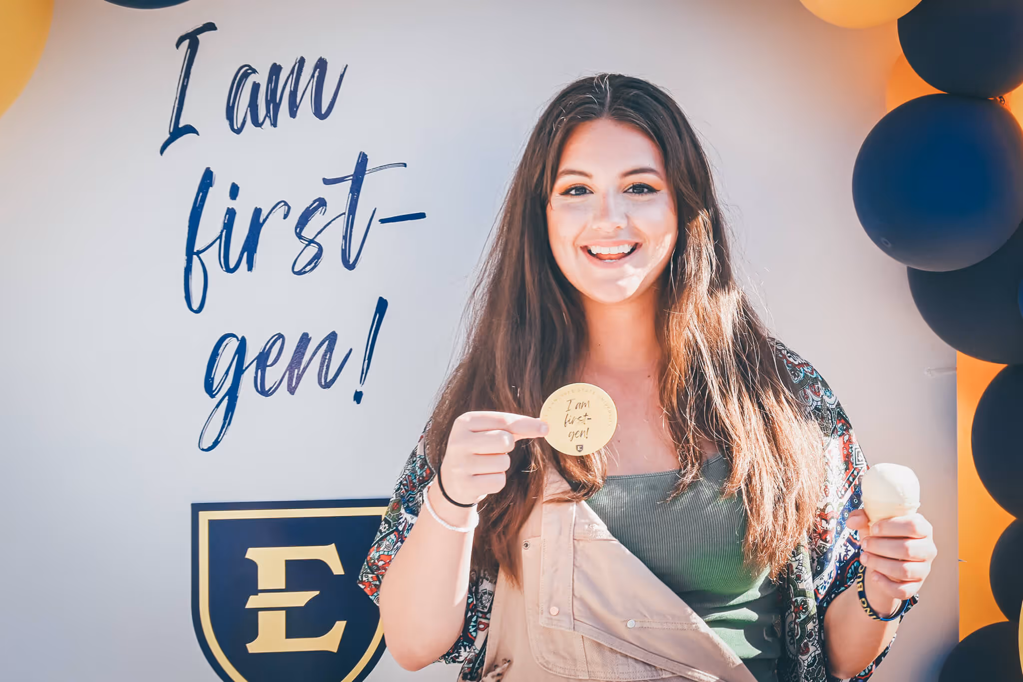 Student smiling while holidng a sticker that says "I am first-gen!" and an ice cream cone.