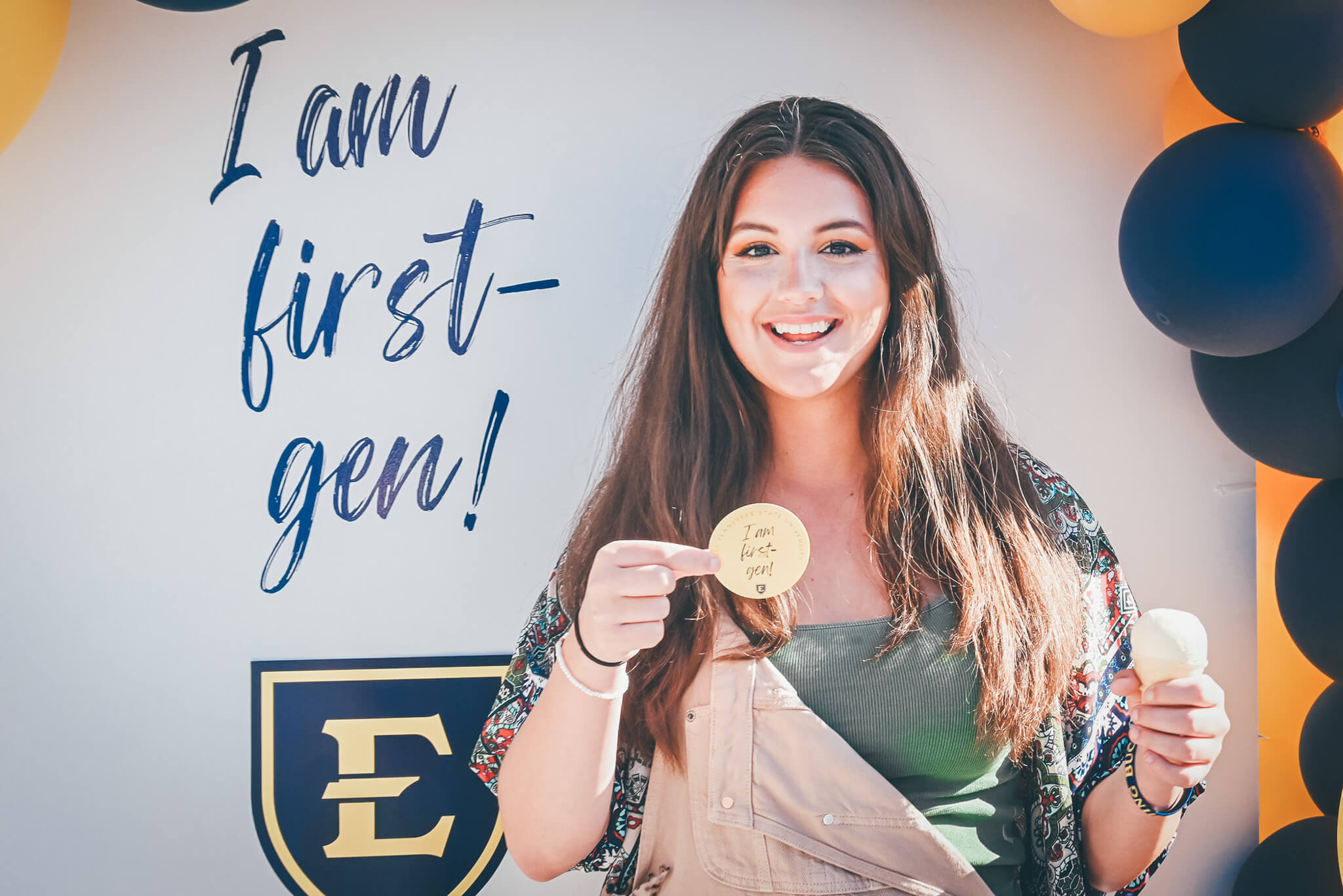 Student smiling while holidng a sticker that says "I am first-gen!" and an ice cream cone.