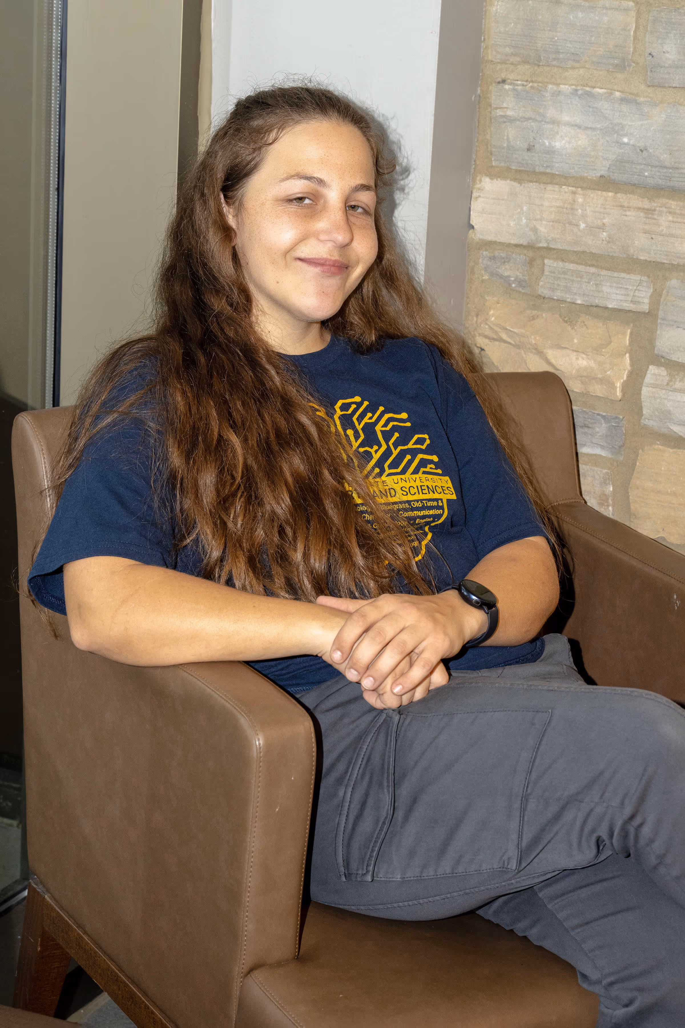 Sidney sits on a tan chair in the Culp Center next to a window, legs crossed and arms clasped together as she looks at the camera. 
