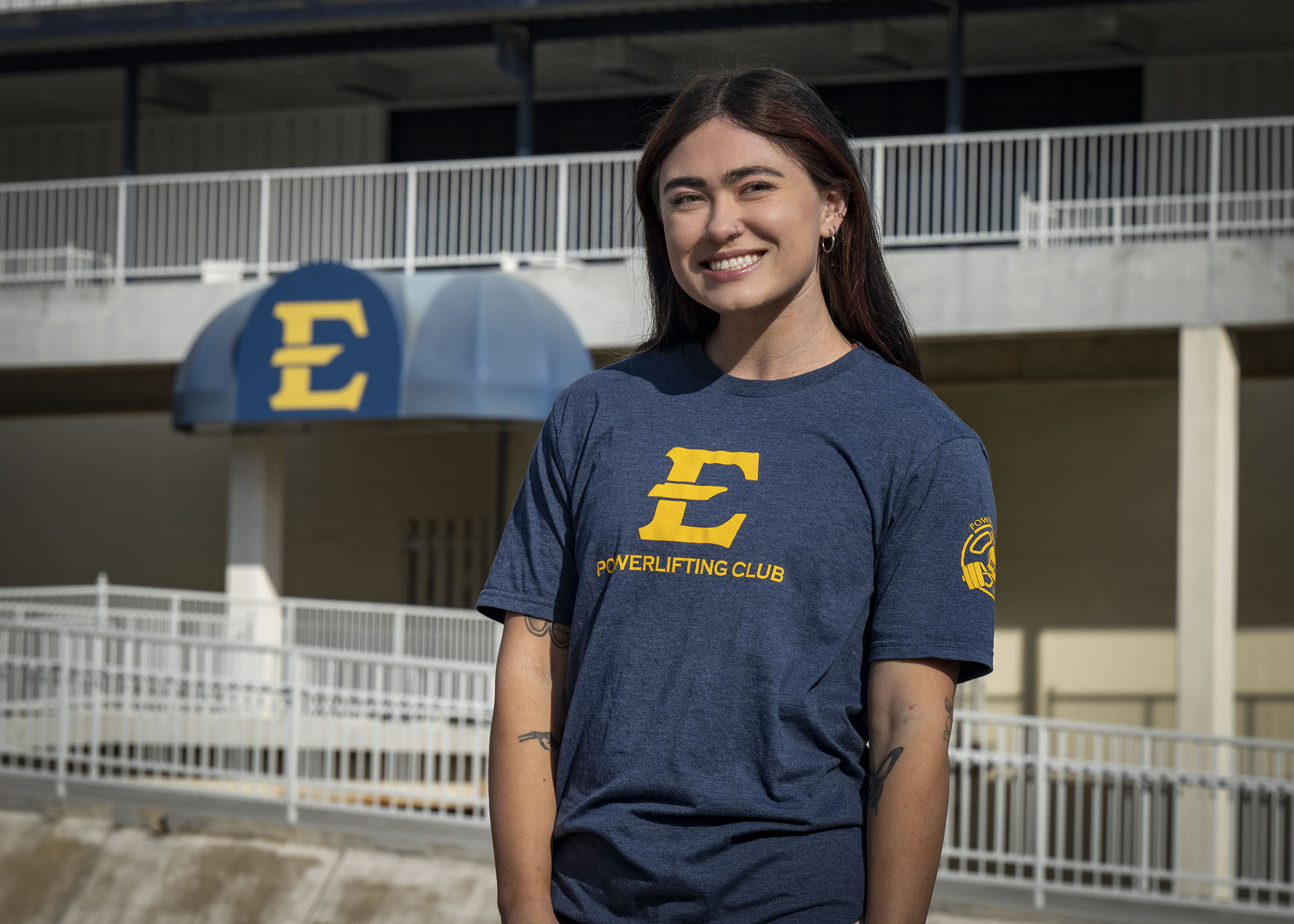 Hannah Burkhart smiling, wearing an ETSU powerlifting shirt with the Ballad Health Athletic Center ramps and doors behind her. 