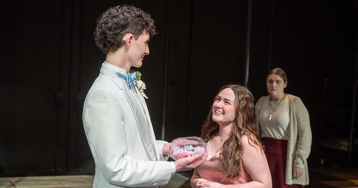 Student actors perform a scene onstage as one character presents a small box while another looks on in the background.