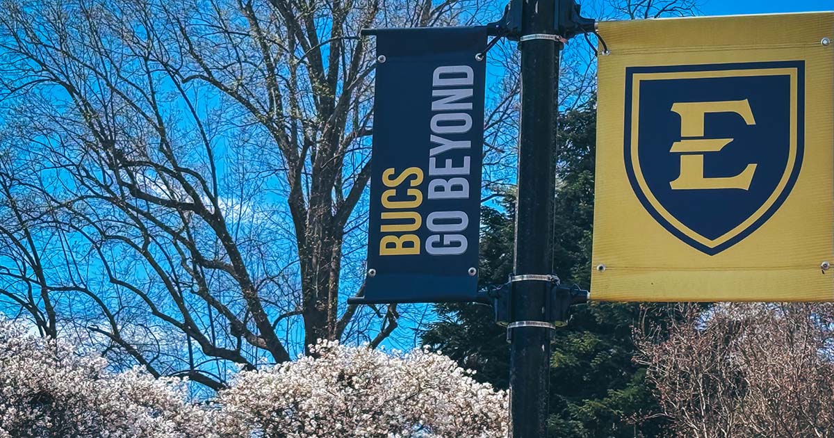 ETSU campus banners reading “Bucs Go Beyond” hang on a lamppost beside blooming trees under a blue sky.