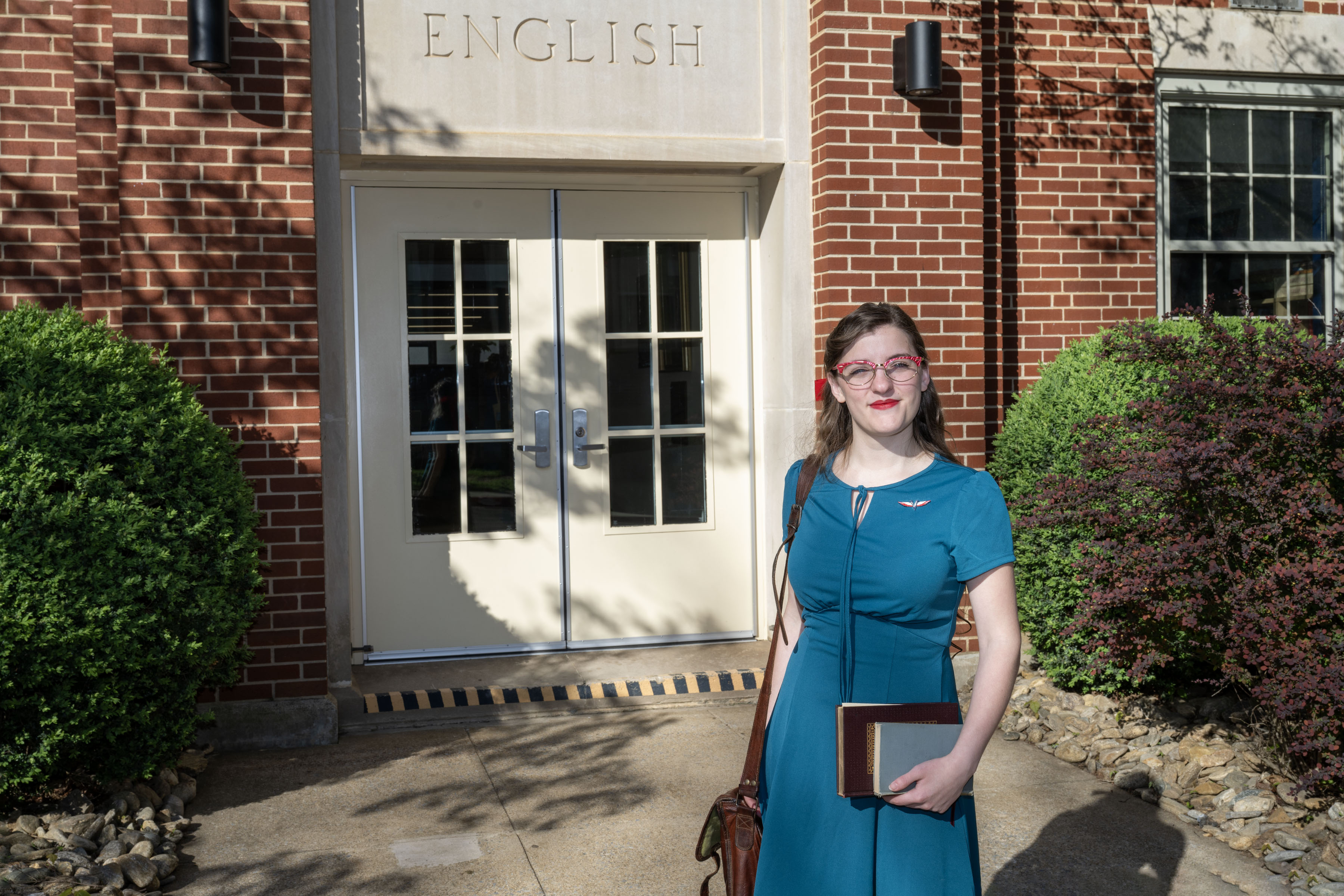 Emerson stands at the entrance of Burleson Hall, holding her bag with one arm and books in the other.