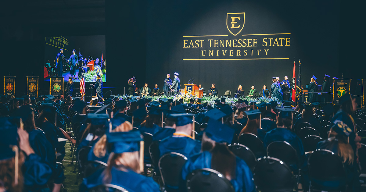 Graduates in caps and gowns sit and stand together inside the Mini-Dome during a graduation ceremony, facing the stage as they celebrate the occasion.