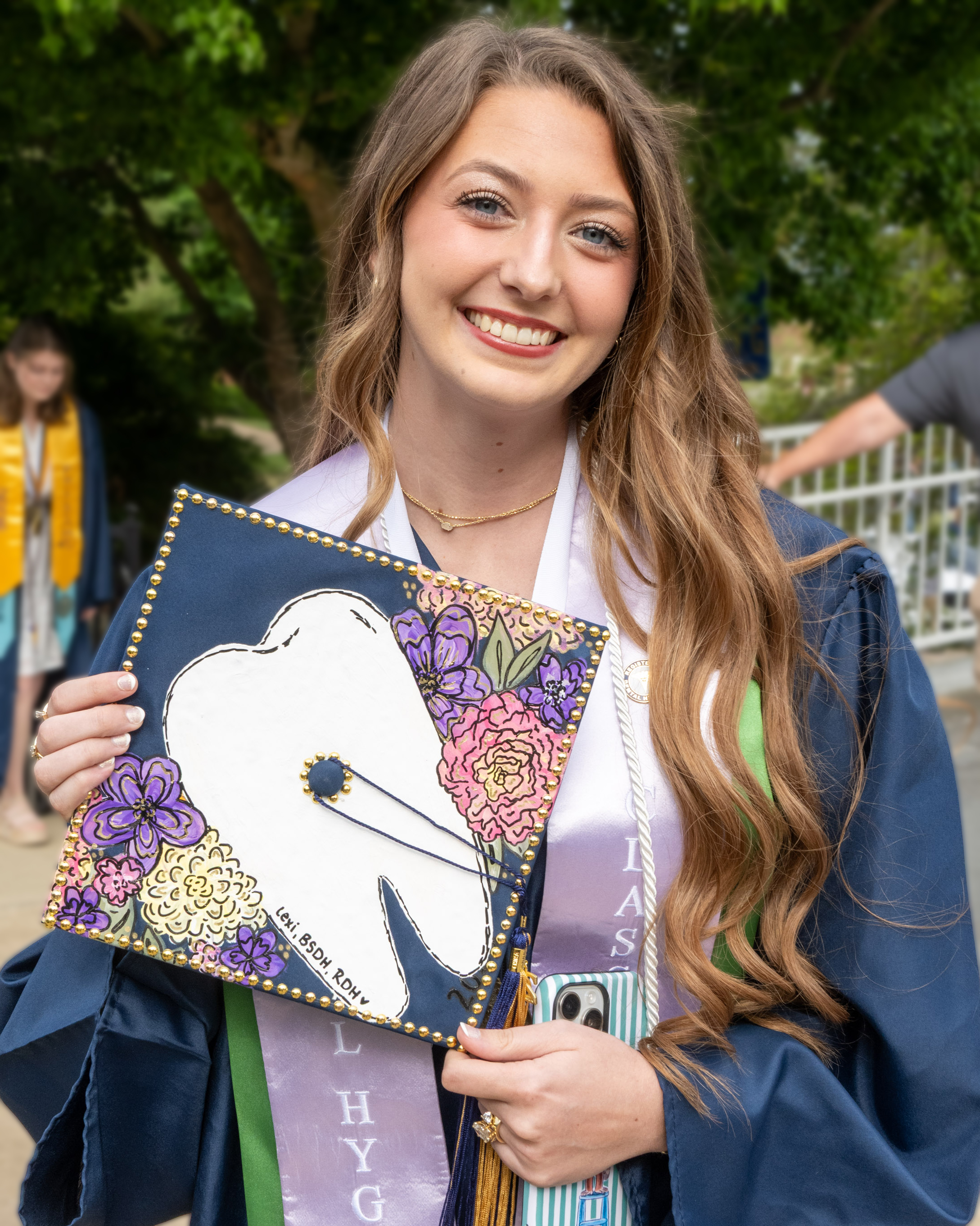 A graduate shows off her decorated grad cap, which has a tooth on it. Her graduation stole indicates she is graduating from ETSU's dental hygiene program.