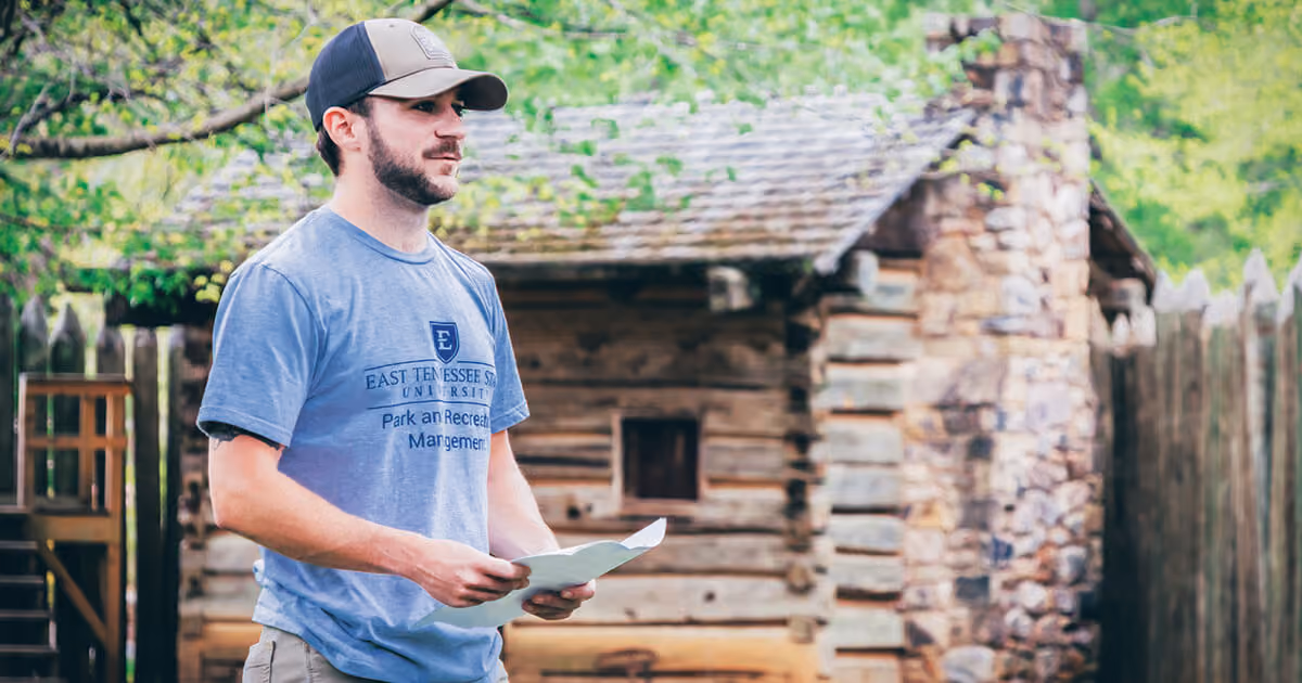 An ETSU Parks and Recreation Management student standing outside one of the cabins at Sycamore Shoals State Historic Site