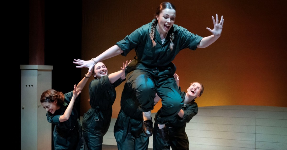 A performer smiles as she's pushed by their castmates, who are also smiling in the background.