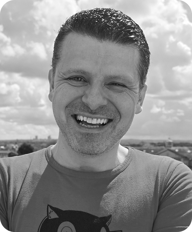 Black and white image of Matheus Guimaraes, leaning over looking into the camera with a wide smile.