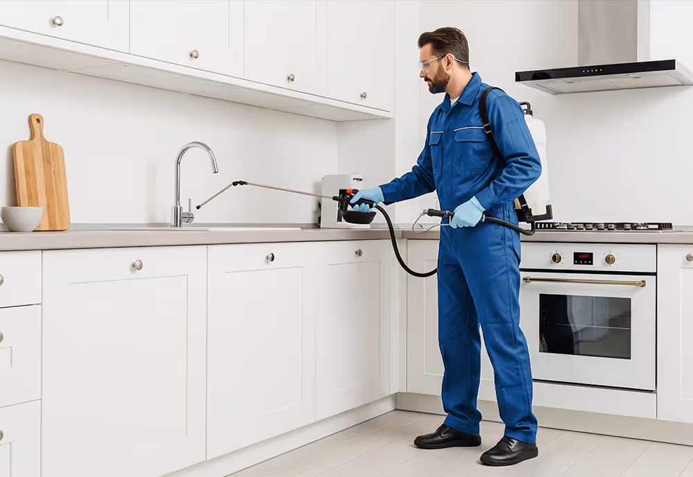 Man in blue uniform and gloves spraying disinfectant around a kitchen sink in a modern white kitchen.
