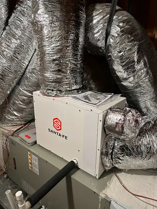 White Santa Fe air ventilation unit connected to large silver insulated flexible ducts in an attic space.