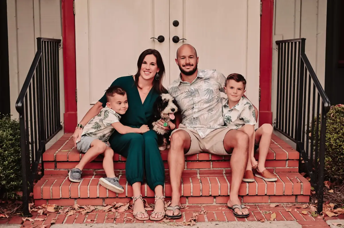 Smiling family of four with a dog sitting on brick steps in front of a white door, parents in the center and children on each side.