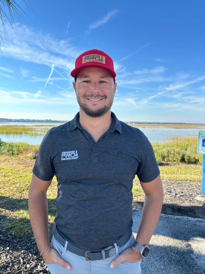 Smiling man wearing a red Temperature Pro cap and dark gray polo shirt standing outdoors near a wetland under a clear blue sky.