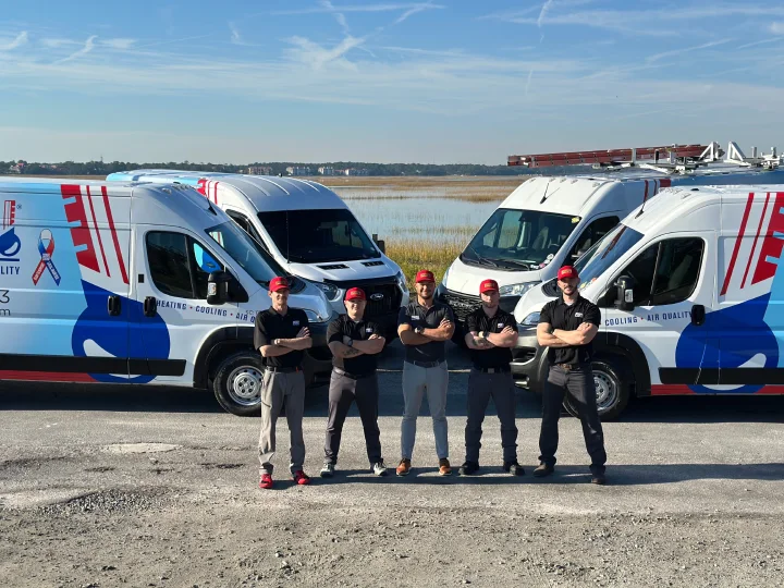 Five technicians wearing black shirts and red hats standing with arms crossed in front of four white service vans by a waterfront.
