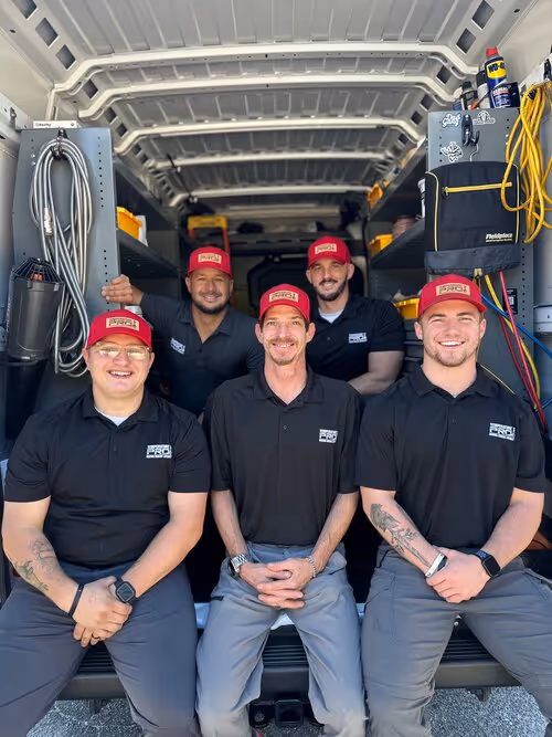 Five men wearing black polo shirts and red hats with logos, sitting and standing inside the back of a work van filled with tools and equipment.