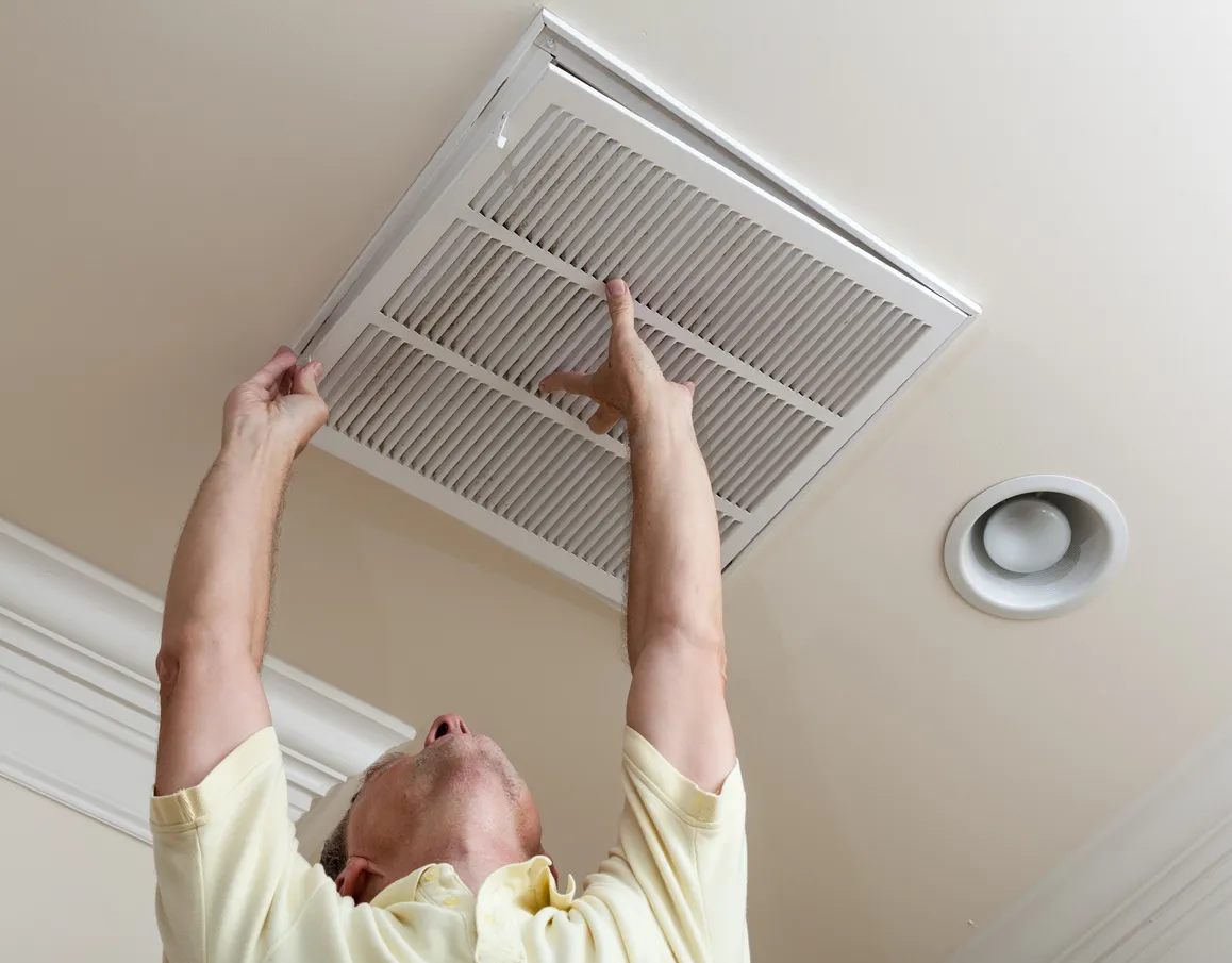 A man replaces a large, pleated white air filter in a ceiling vent