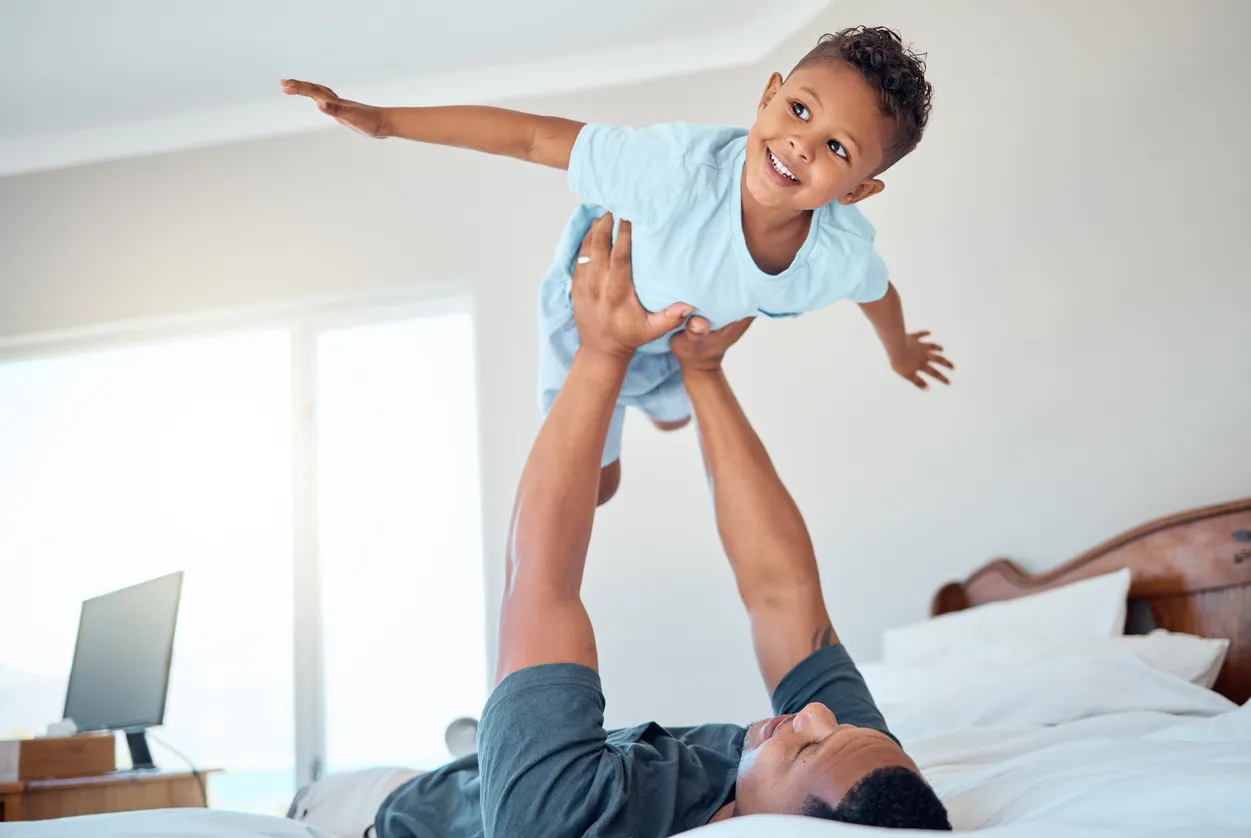 A father lies on a bed, holding his smiling young son up above him in the air