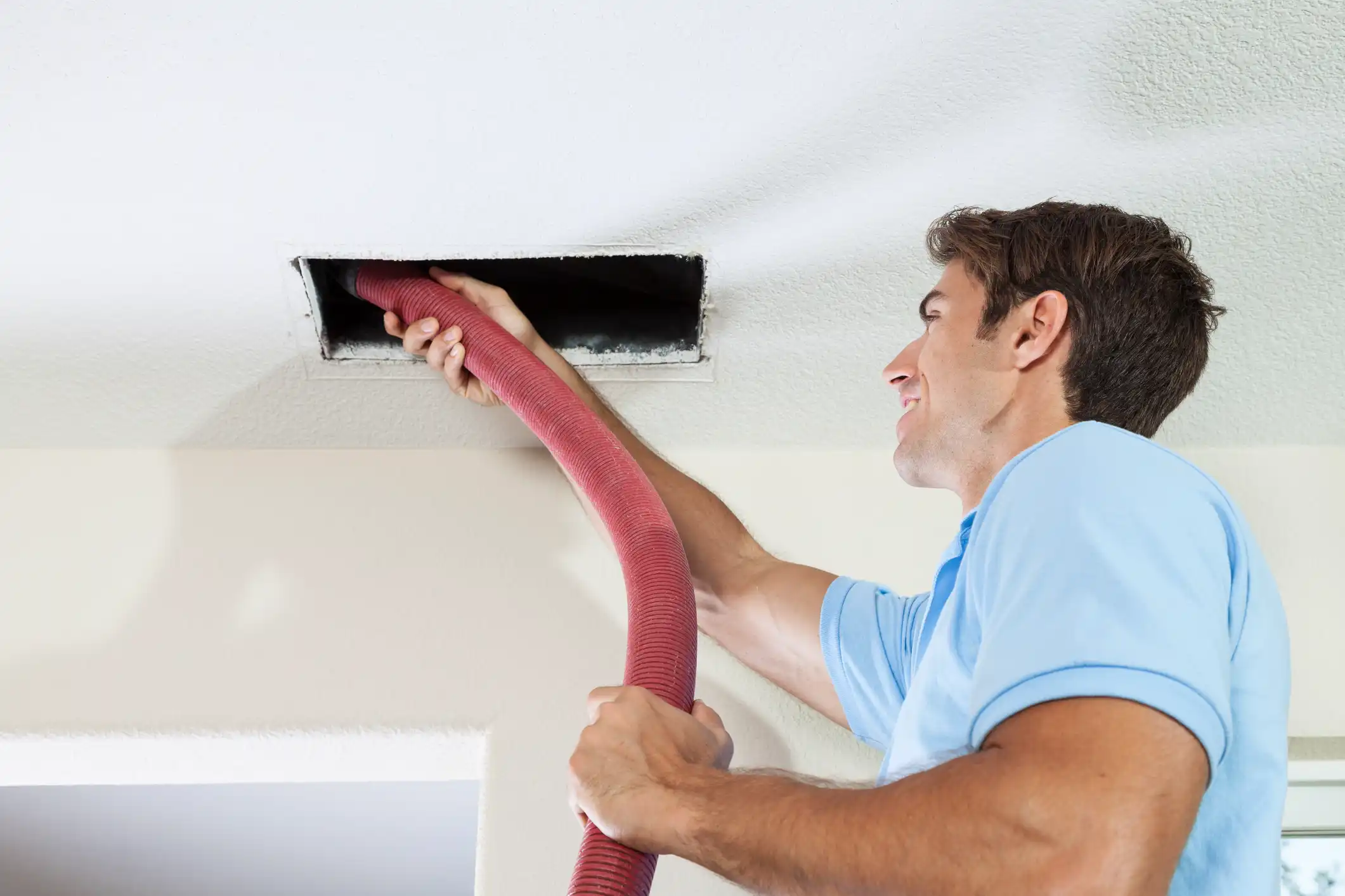  A male technician is inserting a red vacuum hose into a ceiling air duct to clean it.