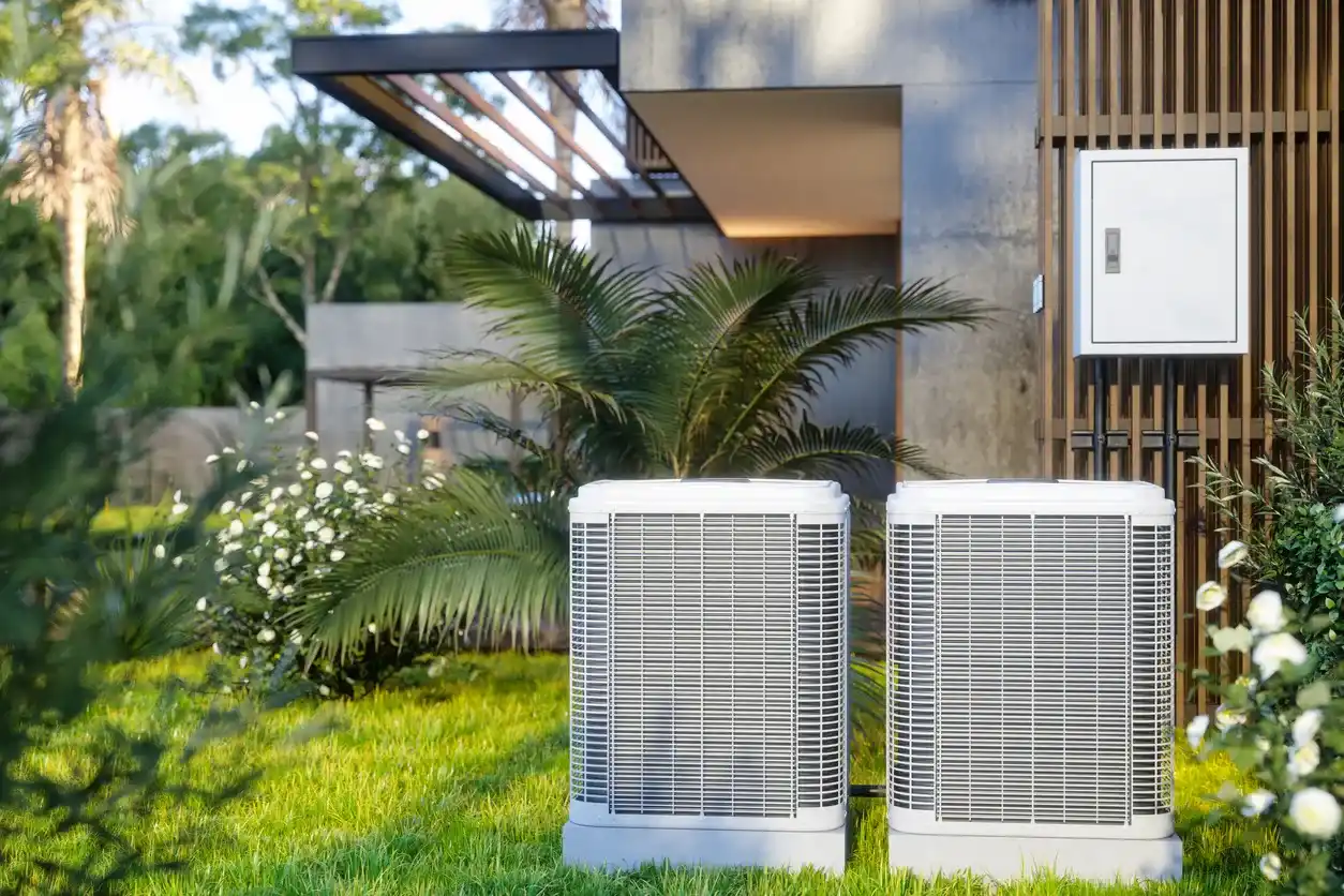 Two identical, modern white heat pump units are installed on a lush green lawn next to a contemporary house with a garden and palm trees.