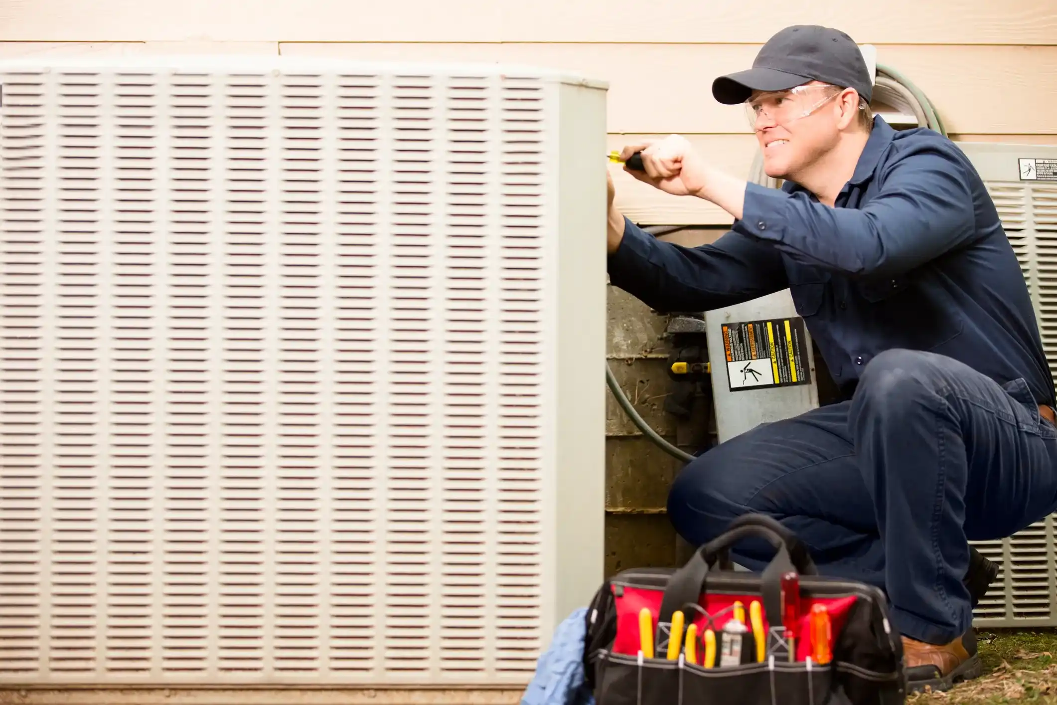 A technician wearing safety glasses and a dark uniform is repairing or servicing a large outdoor HVAC unit with a screwdriver.