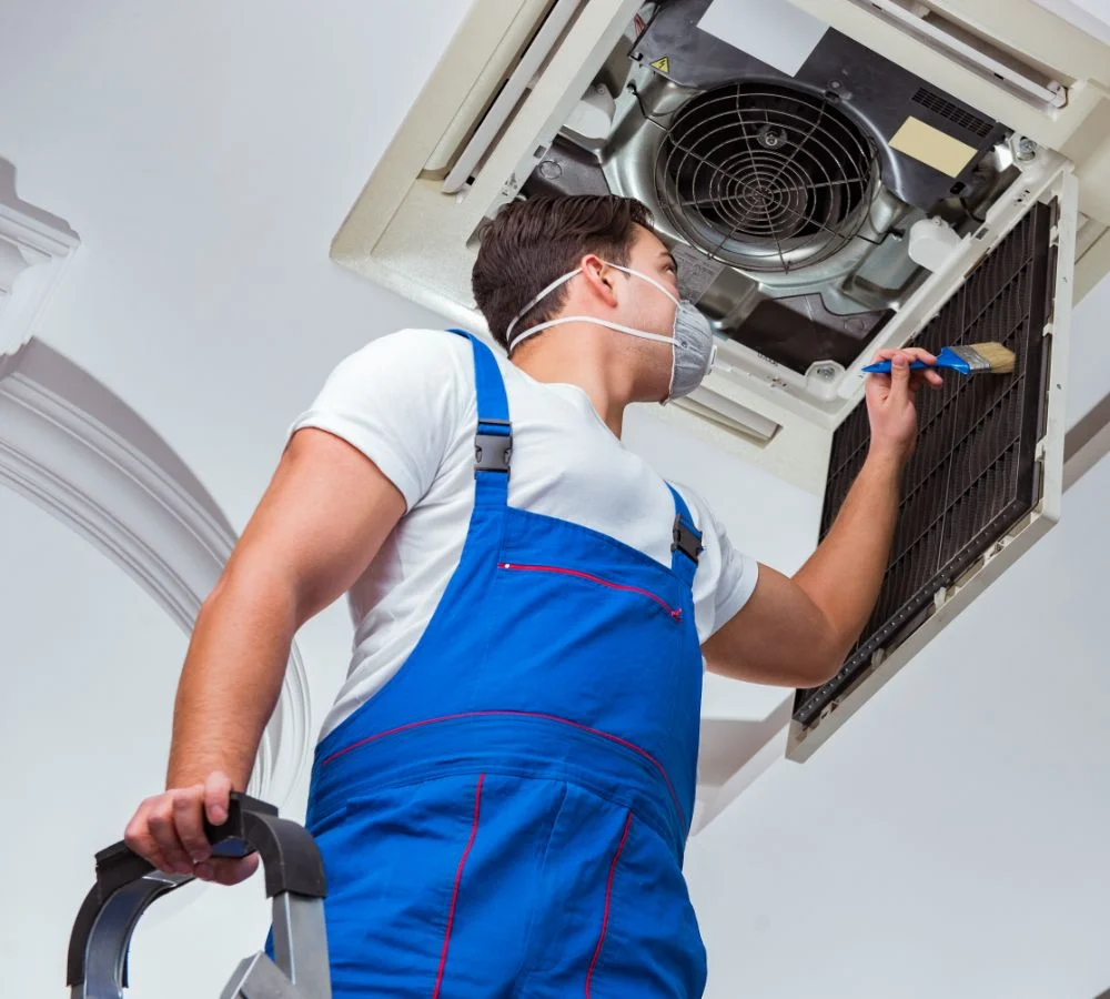 A  service technician in a blue jumpsuit and a face mask is cleaning the inside of a ceiling-mounted HVAC or air conditioning unit with a brush.