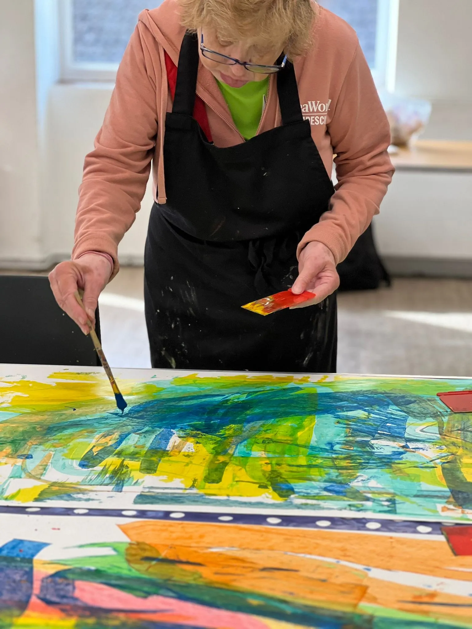 Photo of an artist working on a canvas. They are wearing a pink top and a painting apron. 