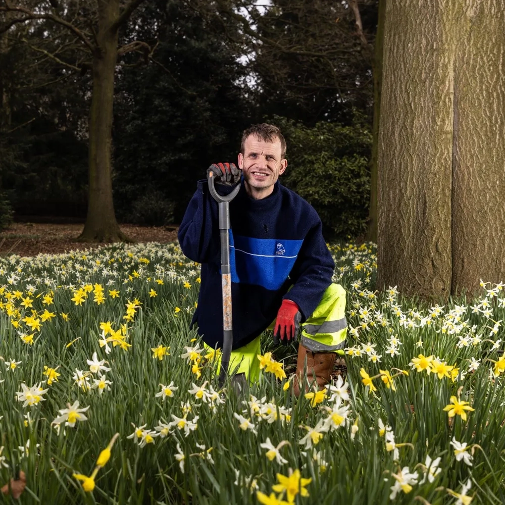 Photo of an ArtWorks artist in outdoor clothes gardening as part of ArtWorks regular volunteer gardening project. The artist has short light brown hair and is wearing a blue jacket and red gardening gloves. They are holding a gardening fork and smiling. They are in a field of daffodils next to a tree.