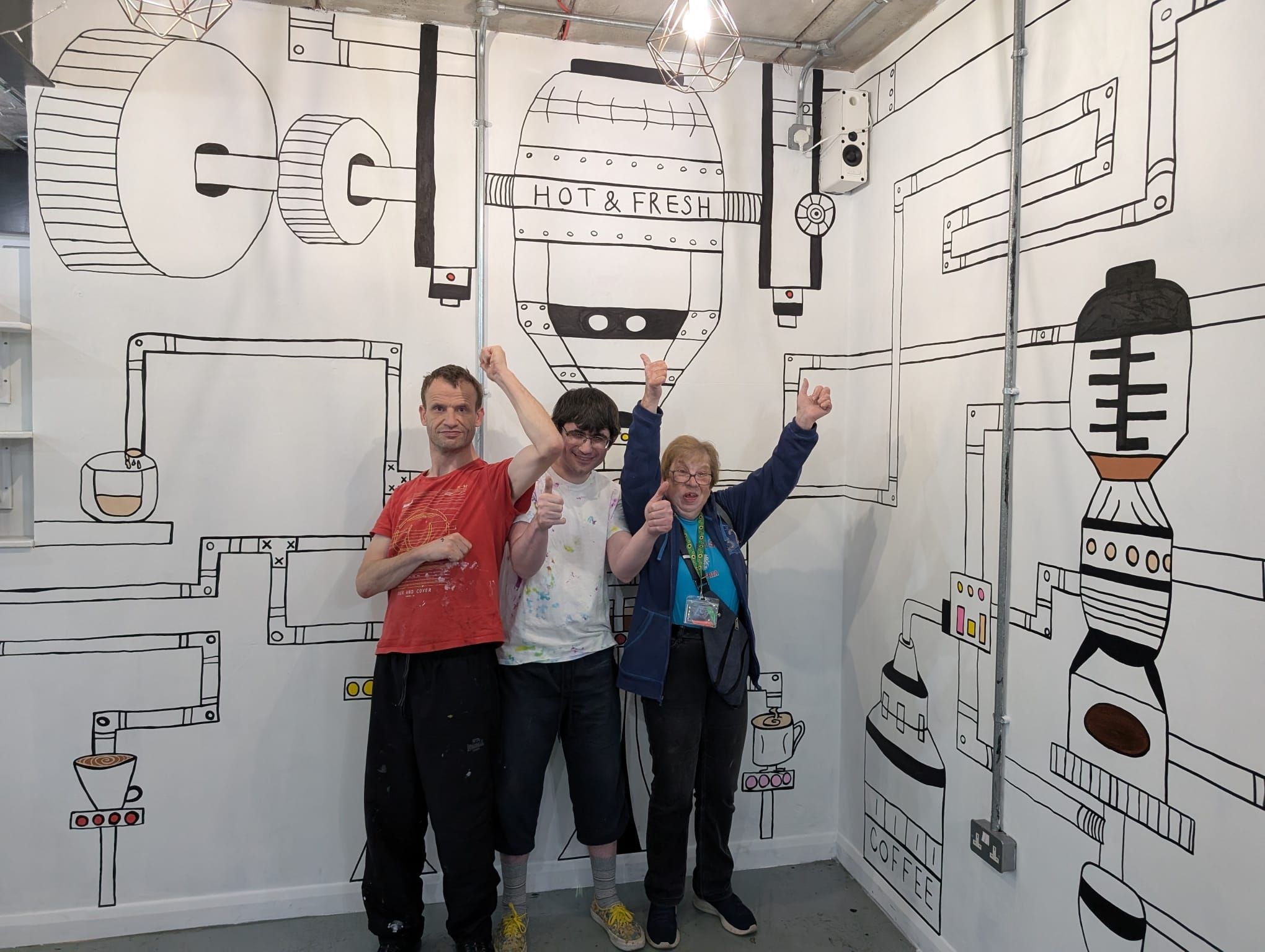 Photo of three ArtWorks artists celebrating the opening of a mural for Theatre Deli coffee shop in Sheffield in 2025. The artists are standing in front of two large white walls decorated with a mural showing how coffee is made. 