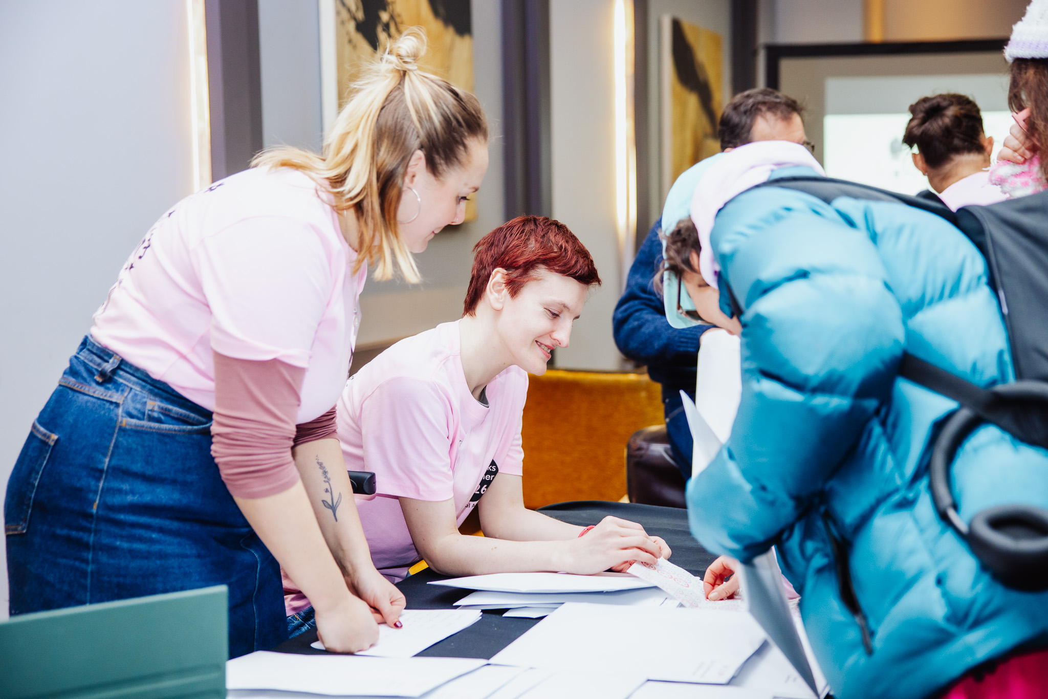 Photo of an ArtWorks artist volunteering at the launch event for ArtWorks Together International Festival 2026. The artist helped visitors when they arrived at the event and gave out information to support their visit. The artist has short red brown hair and is wearing a light pink ArtWorks Together t-shirt. They are speaking with a visitor wearing a blue jacket.