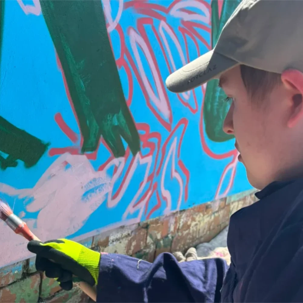 Photo of an ArtWorks artist painting a mural at Maltby in 2026. The artist is wearing a grey baseball cap and painting a wall with blue, green and pink paints. 