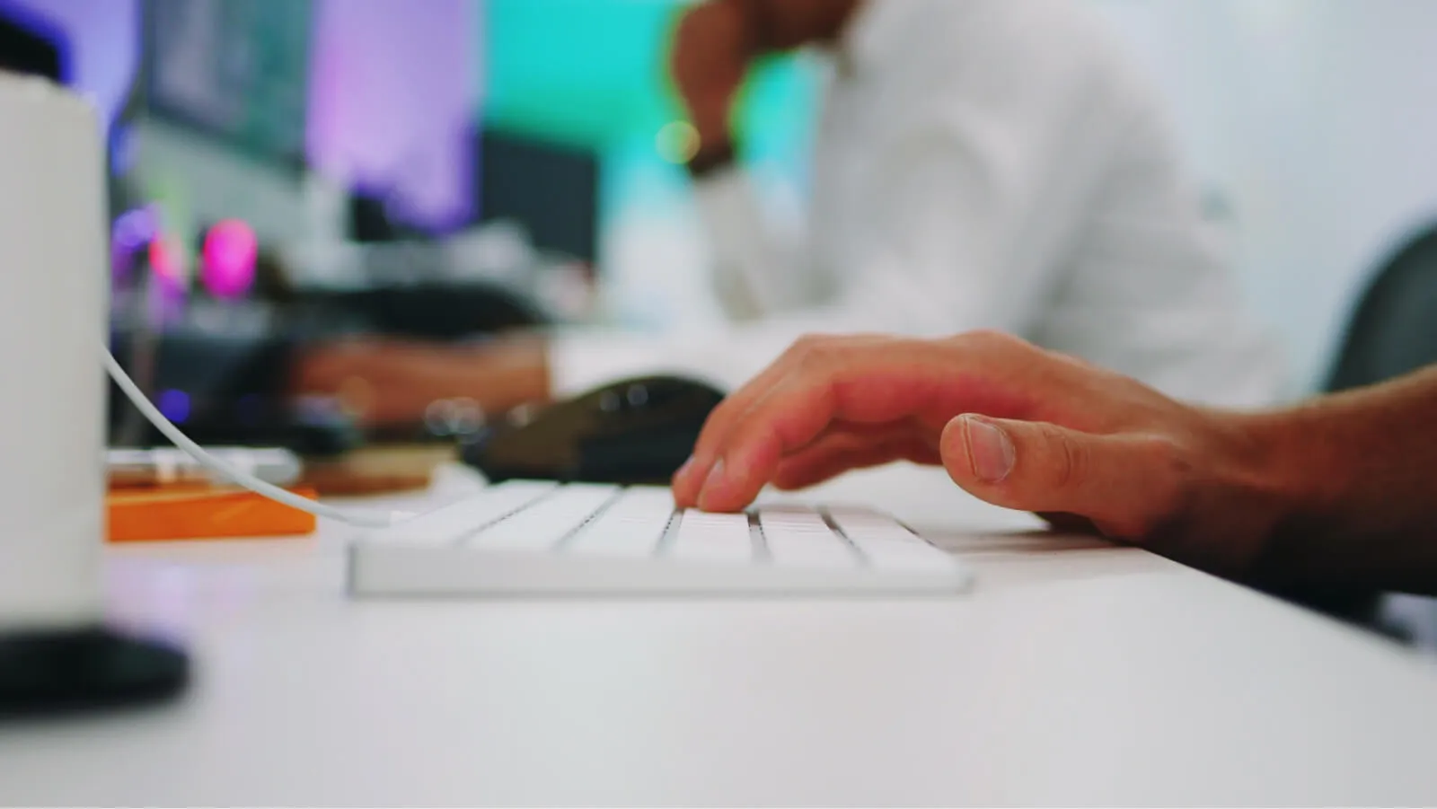 Close-up of a hand typing on a white computer keyboard in an office setting with a blurred person and computer in the background.