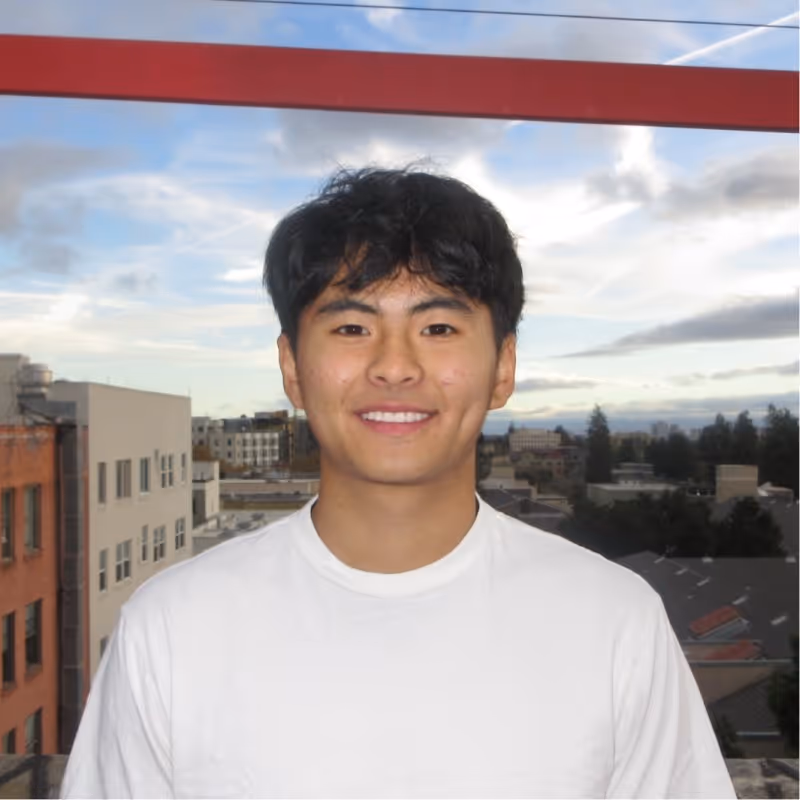 Young man with dark hair wearing a white shirt smiling with city buildings and cloudy sky behind him.