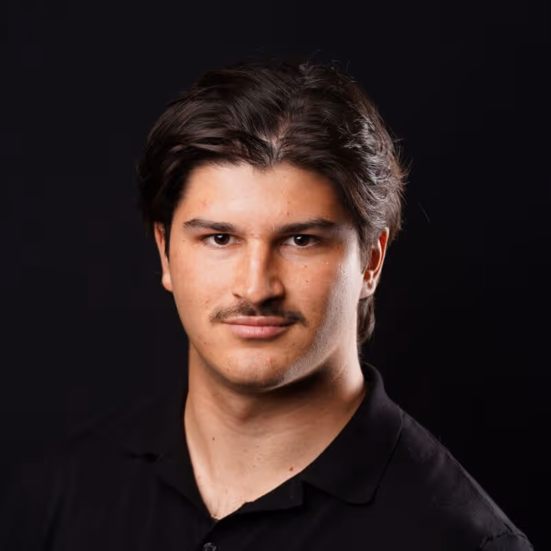 Portrait of a young man with dark hair and a mustache wearing a black collared shirt.
