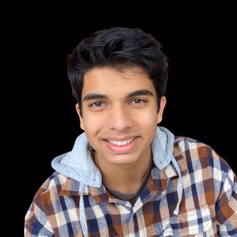 Young man with dark hair smiling, wearing a brown and blue plaid shirt with gray hoodie inside.