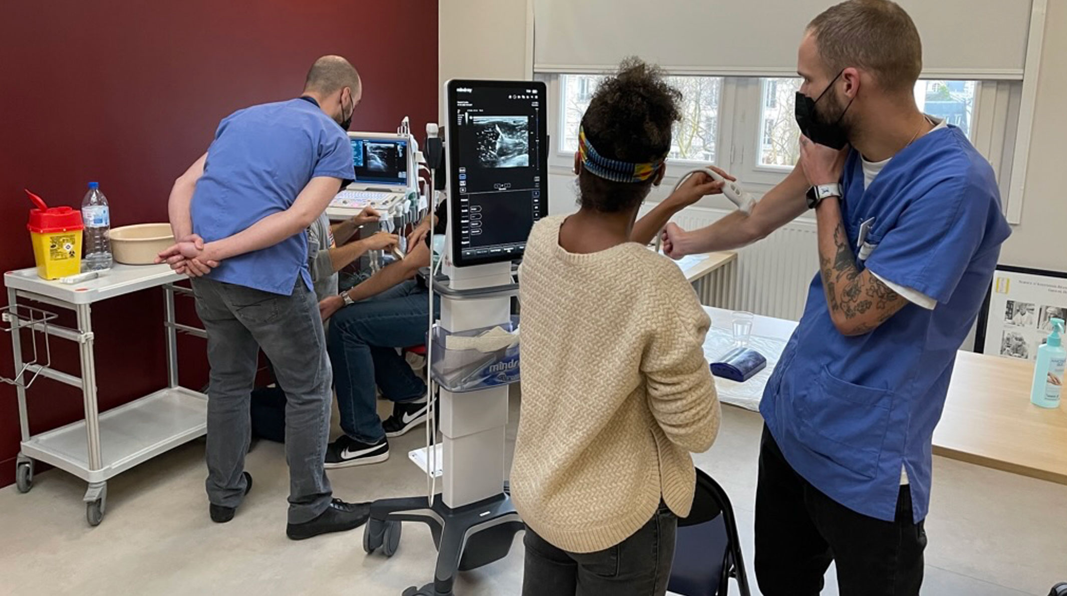 A medical consultation in a clinic where a doctor and a nurse are discussing ultrasound results with a patient, with ultrasound images displayed on multiple screens.