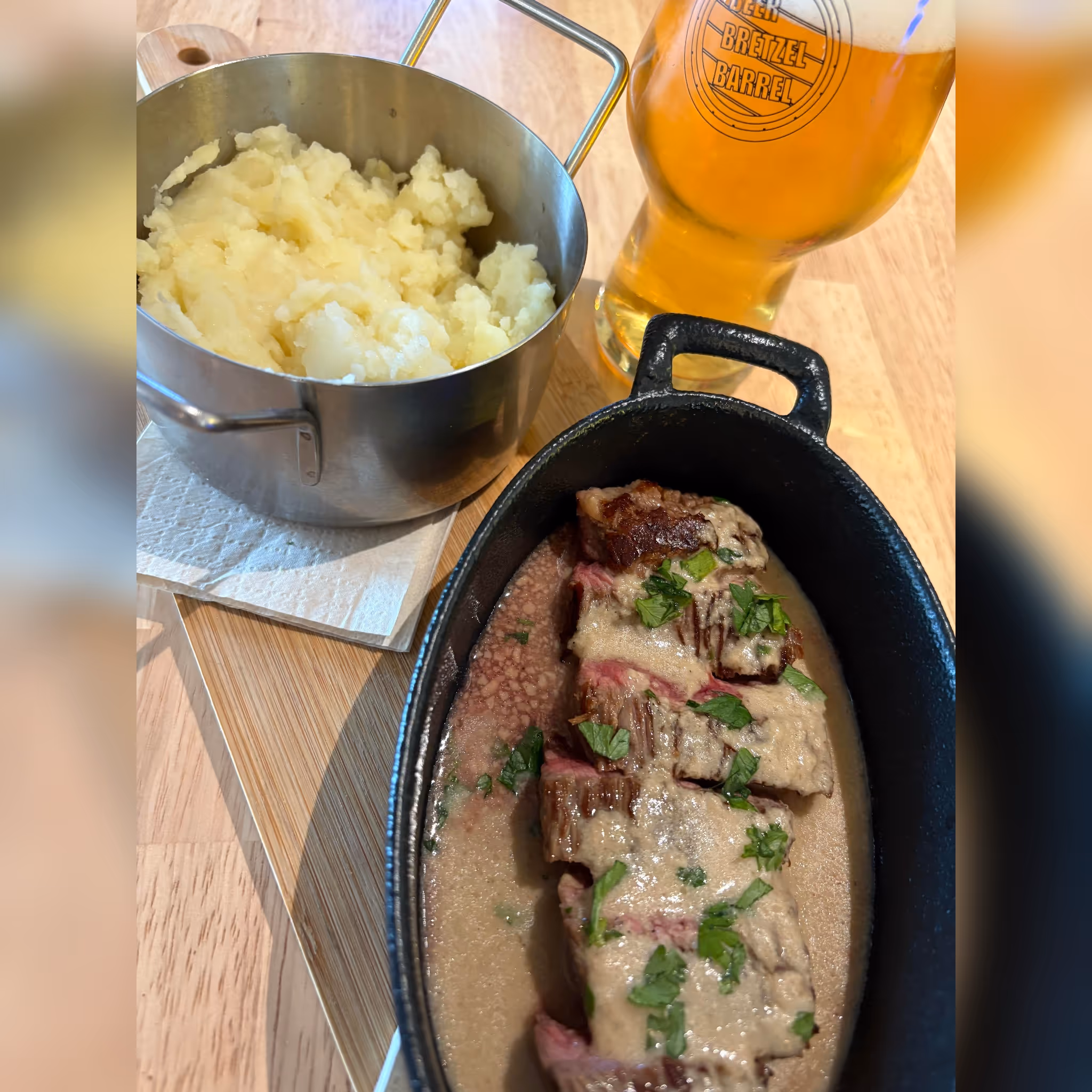 Bifteck avec sauce et persil dans une cocotte noire, purée de pommes de terre dans une casserole en métal et une bière blonde dans un verre estampillé.