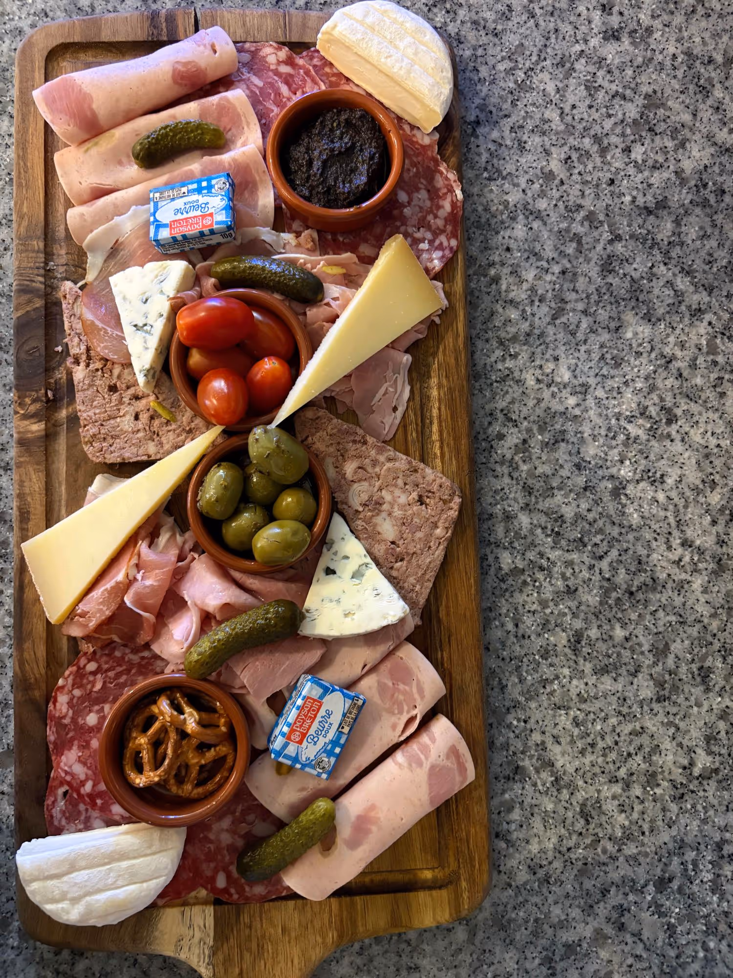 Planche apéritive avec assortiment de charcuterie, fromages, olives, tomates cerises, cornichons, petits bretzels et terrine, présentée sur une planche en bois.