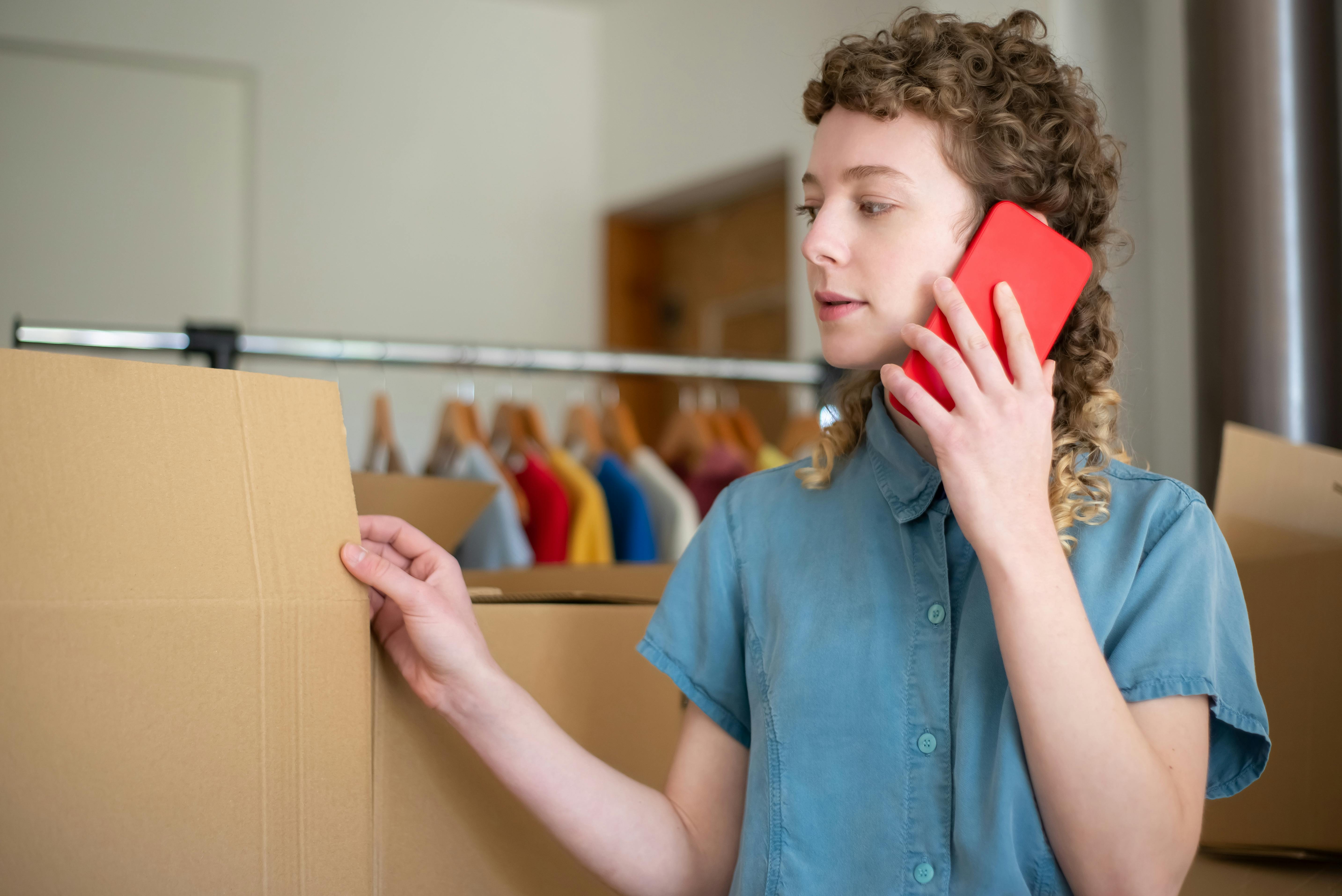 Young woman in blue shirt using phone while packing cardboard boxes indoors.