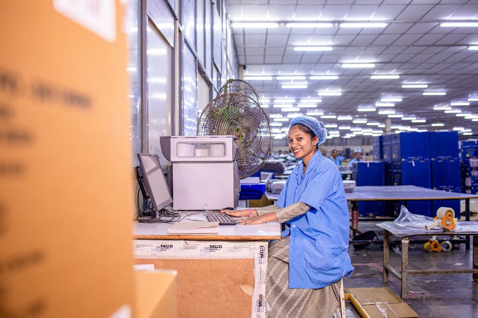 A cheerful female worker at a computer station in a busy warehouse environment.