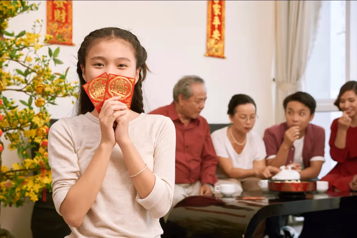 Young girl holding red envelopes in the foreground with family members.