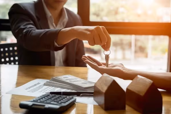Person handing over house keys with money, contract, and calculator on table.