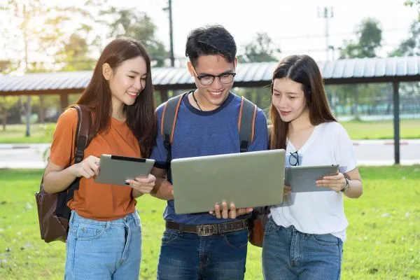 Three students standing outdoors sharing a laptop and tablets.