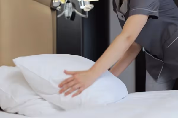A hotel housekeeper in a grey uniform smoothing out a white pillow on a neatly made bed.