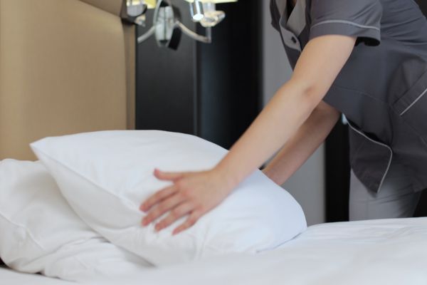 A hotel housekeeper in a grey uniform smoothing out a white pillow on a neatly made bed.