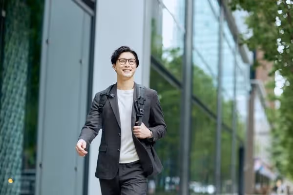 A smiling man in a grey suit and glasses walking outdoors in a modern city business district.