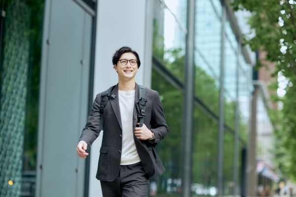 A smiling man in a grey suit and glasses walking outdoors in a modern city business district.