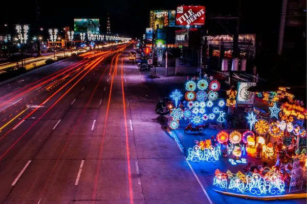 A long-exposure night view of a busy highway with light trails from cars and a colorful display of traditional Filipino Christmas lanterns (parols) on the roadside.