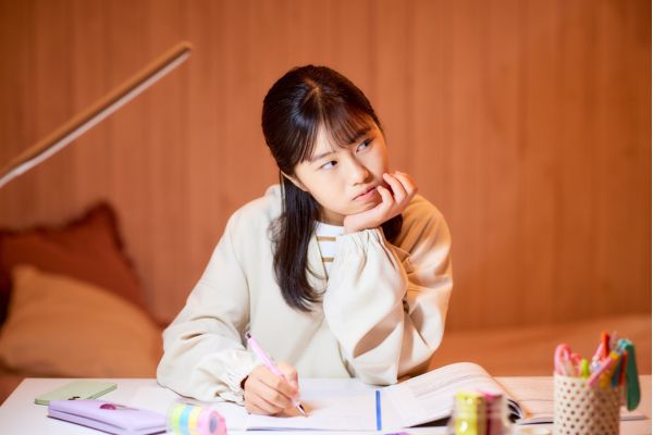A young girl sits at a desk in her condo for students, resting her chin on her hand and looking thoughtful while writing in a notebook. 